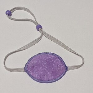 May include: A purple eye patch with a white elastic strap and purple beads. The patch has a swirl pattern.