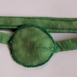 May include: A green eye patch with a green elastic band. The eye patch is made of soft fabric and has a stitched edge.