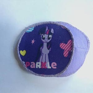 May include: Purple eye patch featuring a cartoon pony with the word "Sparkle" in pink and white. The patch has a purple border and includes a pink star, a yellow heart, and a pink and white striped shape.