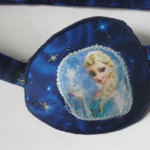 May include: Blue eye patch with a white and blue image of a girl with blonde hair and a blue dress. The background of the eye patch is a dark blue with gold glitter stars.