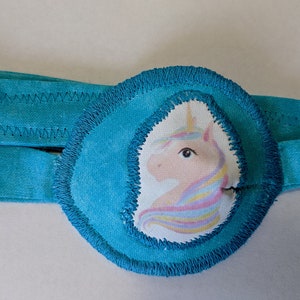 May include: A turquoise eye patch with a unicorn design. The patch has a soft, plush texture and is designed for children.