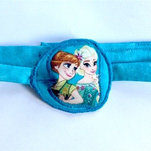 May include: A blue eye patch with a round, padded center featuring an image of two girls with long hair. The girls are wearing dresses and are looking in different directions. The eye patch is made of soft fabric and has a blue elastic band.