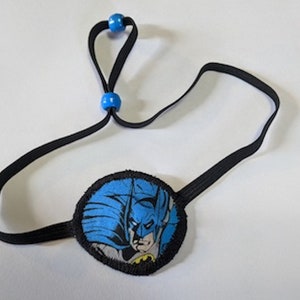 May include: A black eye patch with a Batman graphic. The patch is round with a blue and black design. It has a black elastic strap with blue beads for adjustment. The eye patch is designed for cosplay or costume use.