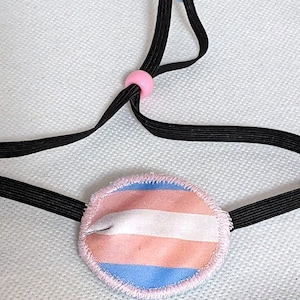 May include: A pink, white, and blue eye patch with a transgender pride flag design. The patch is attached to a black elastic strap with pink and blue beads.