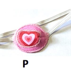May include: Pink eye patch with a pink and white heart design. The eye patch is attached to a white elastic strap.