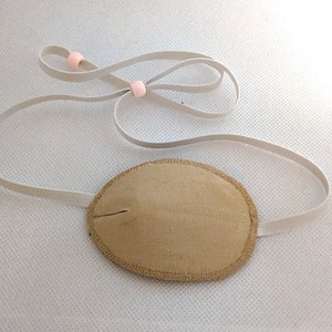May include: A beige eye patch with a white elastic strap and pink plastic adjusters. The eye patch is made of fabric and has a stitched edge.