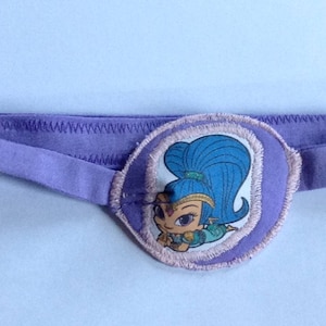 May include: Purple eye patch with an embroidered image of a cartoon character with blue hair and a purple and green outfit. The character is wearing a crown and has a sparkly design.