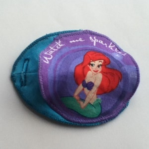 May include: A teal eye patch with a circular patch featuring Ariel from The Little Mermaid. The circular patch has the text "Watch me sparkle". Ariel has red hair, a purple seashell bra, and a green tail.