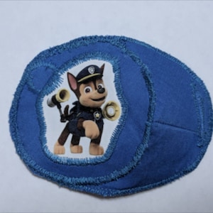 以下が含まれることがあります： パウ・パトロールのキャラクター、チェイスが描かれた青色の布製パッチ。茶色とベージュの子犬であるチェイスは、警察官の制服と帽子を着用しています。パッチは円形で、縁がほつれています。