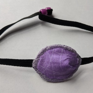 May include: Purple eye patch with an elastic strap. The patch is made of soft fabric and has a sewn edge.