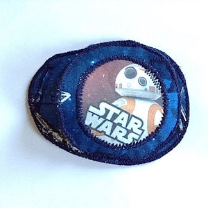 May include: A blue fabric patch featuring a white and orange Star Wars character, BB-8, in the centre. The patch has a dark blue border and the words "Star Wars" are printed in white.