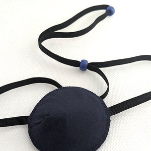 May include: A navy blue eye patch with a black elastic strap and blue adjustment beads. The circular patch is made of fabric and is designed to cover one eye. The elastic strap is adjustable for a comfortable fit.