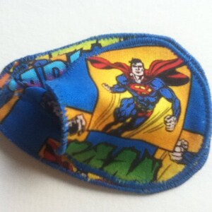 Può includere: Una benda sull'occhio blu con un disegno di Superman. La toppa ha un bordo blu e un design in stile fumetto con Superman in volo, che indossa il suo mantello rosso e il suo costume blu. Lo sfondo è giallo e blu.