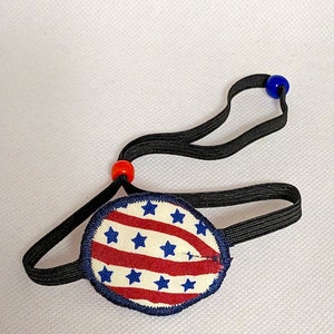 May include: A red, white, and blue eye patch with stars and stripes. The eye patch is attached to a black elastic strap with red and blue beads.