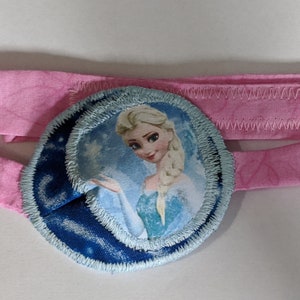 Può includere: Un cerotto per l'occhio rosa con un cerotto in tessuto blu e bianco con un'immagine di Elsa di Frozen. Il cerotto è cucito su una fascia rosa.