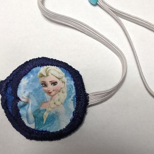 Op de afbeelding: Een blauw en wit ooglapje met een afbeelding van Elsa uit Frozen. Het ooglapje heeft een elastisch bandje met een lichtblauwe kraal.