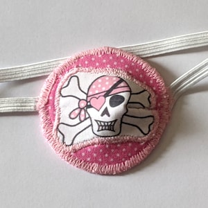 Może przedstawiać: Różowy łatkę na oko z białym wzorem czaszki i skrzyżowanych kości. Czaszka nosi różową chustkę z białym sercem. Łatka na oko jest przymocowana do białej gumki.