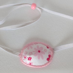 May include: A pink and white eye patch with a floral pattern, attached to a white elastic strap with pink beads.