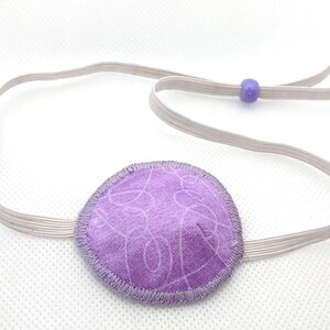 May include: A lavender eye patch with a gray elastic strap and two small purple beads. The eye patch is round with a stitched border and a light purple swirl pattern. The elastic strap is adjustable.