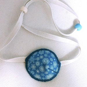 May include: A blue eye patch with a white snowflake pattern. The patch is attached to a white elastic strap with white and blue beads.