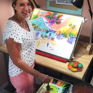 Op de afbeelding: Een vrouw in een wit shirt met stippen en een roze broek schildert een aquarel van een tropische sc&egrave;ne. De schildering toont een blauw gebouw met een palmboom ervoor. De vrouw gebruikt een penseel om verf op het doek aan te brengen. Ze heeft een palet met aquarelverf voor zich.