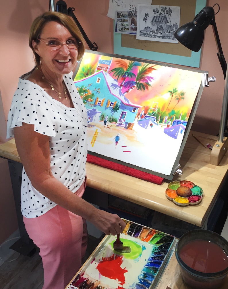 Op de afbeelding: Een vrouw in een wit shirt met stippen en een roze broek schildert een aquarel van een tropische sc&egrave;ne. De schildering toont een blauw gebouw met een palmboom ervoor. De vrouw gebruikt een penseel om verf op het doek aan te brengen. Ze heeft een palet met aquarelverf voor zich.
