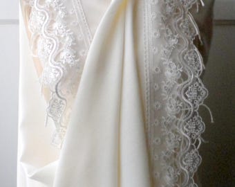 May include: Cream-coloured shawl with detailed white lace edging. The shawl is draped over a mannequin, highlighting its flowing fabric and delicate design. The lace features floral patterns.