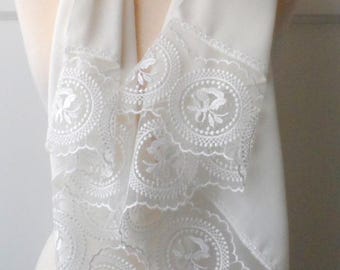 May include: Cream-coloured scarf with detailed white lace edging. The scarf is displayed on a mannequin, highlighting its delicate texture and design. The lace features circular patterns with floral motifs, adding elegance.