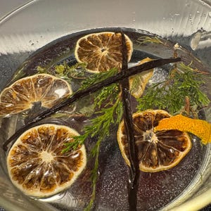 May include: A clear glass bowl filled with water containing three dried citrus slices, a bright orange peel, two dark brown vanilla beans arranged in an X shape, and several sprigs of green evergreen needles submerged in the liquid.