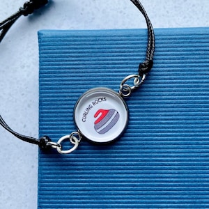 May include: A black string bracelet with a silver charm featuring a curling rock and the text "Curling Rocks".