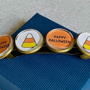 May include: A set of four Halloween-themed hair clips. Each clip features a round, clear glass cabochon with a different design. One clip has a candy corn design, another has the text "Happy Halloween", and the other two have a candy corn design. The clips are attached to a gold-colored metal bar.