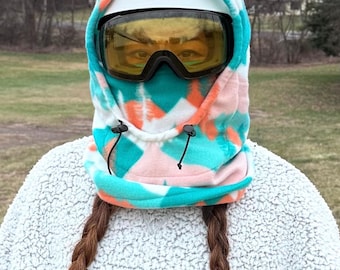 Könnte beinhalten: Eine Balaclava im Batik-Stil in Türkis, Koralle und Weiß, getragen mit einem weißen Helm und gelb getönten Schutzbrillen. Die Sturmhaube hat einen Kordelzug und bedeckt Hals und Kopf. Die Person trägt einen weißen, flauschigen Pullover.