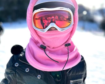 Puede incluir: Una máscara de esquí de forro polar rosa con un casco blanco y gafas de esquí. Las gafas tienen una lente reflectante con un tinte rojo y naranja.