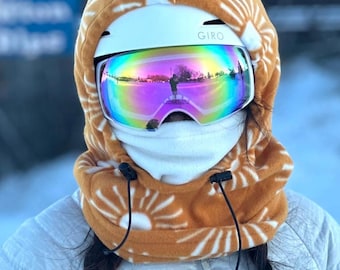May include: A white ski helmet with a brown fleece balaclava with a white sun pattern. The helmet has the word "GIRO" printed on it. The person is wearing iridescent ski goggles.