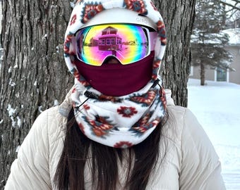 May include: A person wearing a white ski helmet with rainbow-tinted goggles and a burgundy neck gaiter. The helmet and gaiter have a floral pattern. The person is wearing a light beige jacket, standing in front of a tree in a snowy environment.