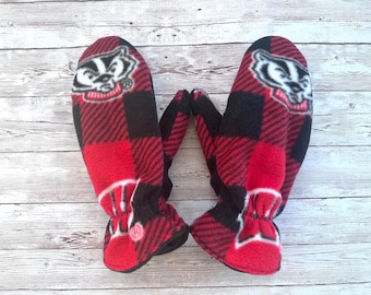 May include: A pair of red and black fleece mittens with a white badger logo. The mittens have a plaid pattern and are lined with fleece.