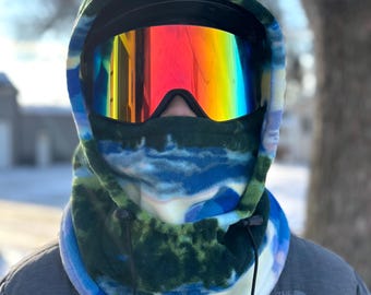 Puede incluir: Un pasamontañas de cuello de forro polar azul, verde y blanco con cierre de cordón. El pasamontañas se lleva sobre la cabeza y la cara de una persona, con unas gafas de esquí de colores arcoíris que cubren los ojos.