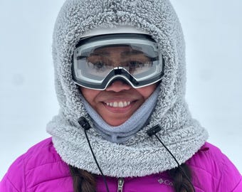May include: A person wearing a gray fleece hooded neck gaiter with black straps and ski goggles. The person is also wearing a pink quilted jacket with a zipper and two braids.