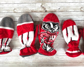 May include: A collection of red and white mittens with various designs. Some mittens feature the letter "W", others display a badger mascot, and some have the word "BADGERS". The mittens are made of a soft, fleece-like material.
