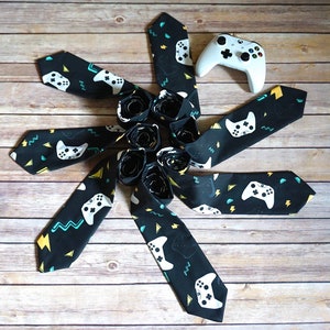 May include: A set of eight black neckties with a white and yellow gamepad pattern. The ties are arranged in a circle around a white video game controller.