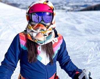 May include: A person wearing a blue and white ski suit with a colorful patterned hood and pink helmet. The person is holding ski poles and is standing on a snowy slope.