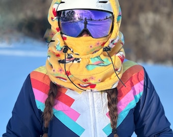 May include: A person wearing a yellow fleece ski mask with a colorful pattern, a white ski helmet with the word "GIRO" on it, and purple ski goggles. The person is wearing a blue ski suit with a rainbow pattern on the chest and a red belt.