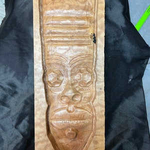 May include: A wooden carving of a face with a detailed expression. The carving is in a rectangular frame and has a light brown color.