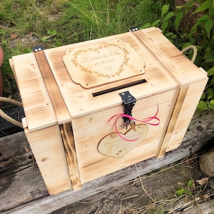 May include: A rectangular wooden box with a hinged lid featuring a slot, engraved with the text Kristin & Eike 16.08.2025. The box has scorched wood accents, a black metal latch, and a heart-shaped wooden tag tied with a pink ribbon.