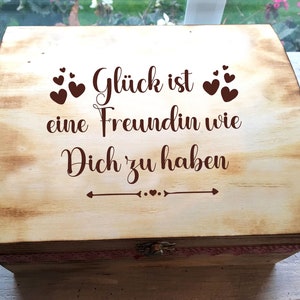 Peut inclure: Une boîte en bois avec une finition vieillie et un ruban rose. Le couvercle porte une inscription brune qui dit "Glück ist eine Freundin wie Dich zu haben" ce qui se traduit par "Le bonheur est d'avoir un ami comme toi".