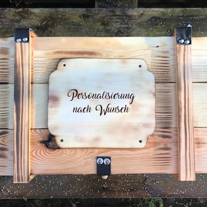 Könnte beinhalten: Eine rechteckige Holzkiste mit heller Naturholzoptik und geflammten Kanten. Auf einer mittigen Holzplatte ist der Text "Personalisierung nach Wunsch" eingraviert. Die Kiste verfügt über schwarze Lederriemen mit Metallnieten und Seilgriffe.