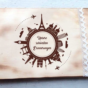 May include: A wooden photo album with a brown background and a design of a world map with famous landmarks. The album has a white lace trim and the text "Unsere sch&ouml;nsten Erinnerungen" is printed on the front.