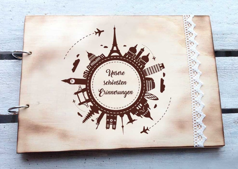 May include: A wooden photo album with a brown background and a design of a world map with famous landmarks. The album has a white lace trim and the text "Unsere sch&ouml;nsten Erinnerungen" is printed on the front.