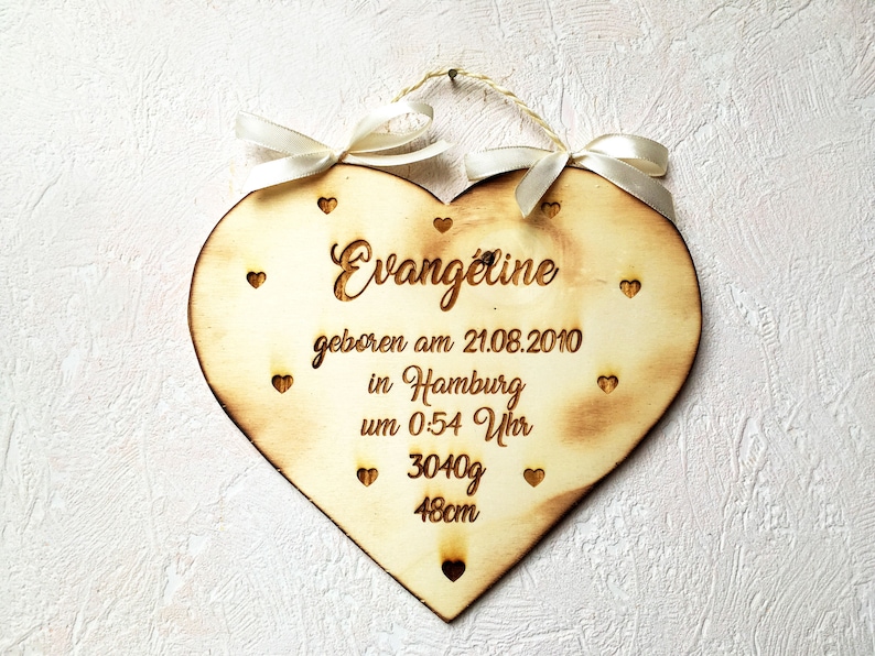 May include: A wooden heart-shaped plaque with the text "Evangeline geboren am 21.08.2010 in Hamburg um 0.54 Uhr 3040g 48cm" engraved on it. The plaque is decorated with small hearts and has a string attached to the top for hanging.