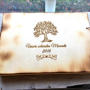 May include: A wooden photo album with a burnt wood finish. The cover features a laser-engraved design of a tree with the text "Unsere sch&ouml;nsten Momente 2018".